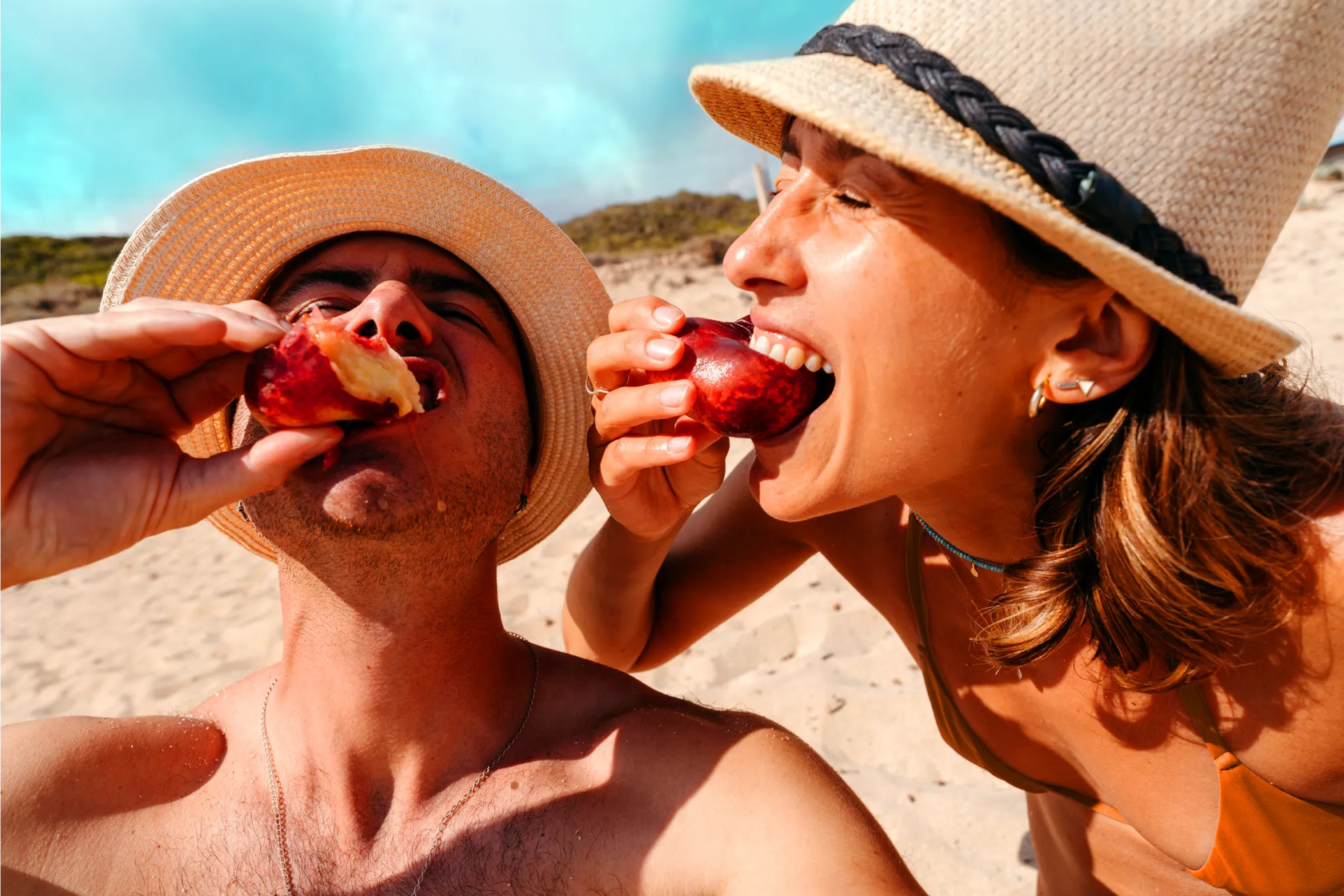 A couple biting into peaches while relaxing on a beach together